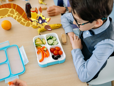 Child sitting in front of lunchbox filled with vegetables, with Sam the giraffe. 