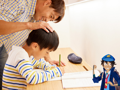 Child sitting at desk filling out worksheet, with his father standing over him looking encouraging. 