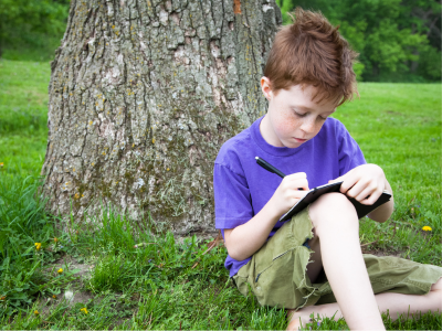 Child sitting under a tree, writing in a journal. 