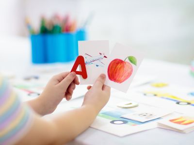 Child holding up a drawing of an aeroplane, a drawing of an apple, and the letter A.